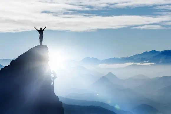 Climber reaching the mountain summit at sunrise with another person cheering on the peak.